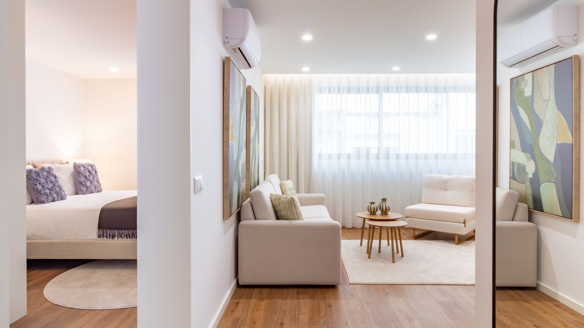 A modern hotel room with a bed, a sofa, a small table with vases, and a large abstract painting on the wall.