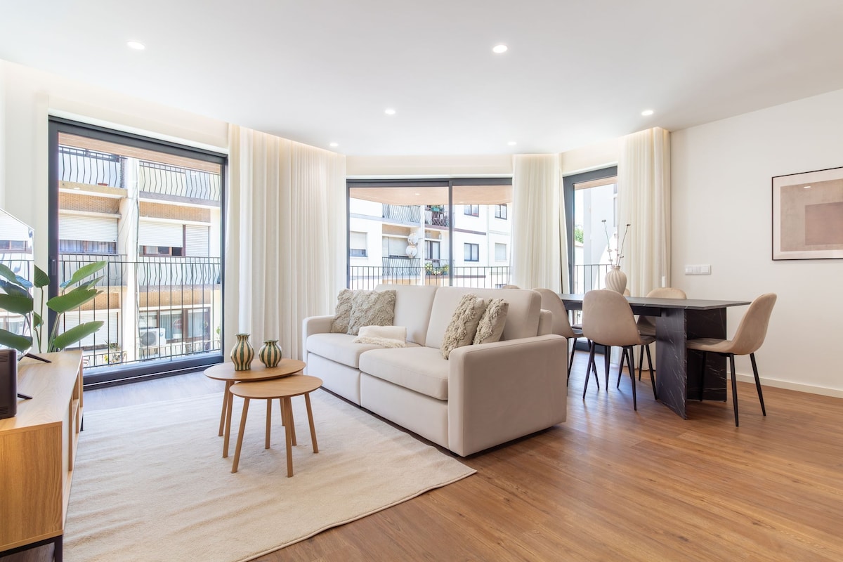 A modern living room with a sofa, a dining table, and chairs. There is a large window with a view of buildings outside.