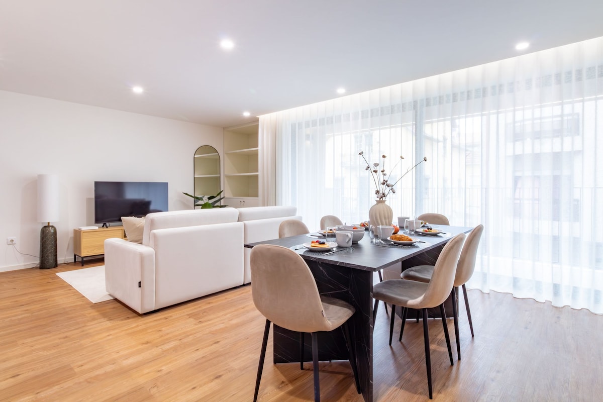A modern living room with a dining area, a sofa, a TV, and a large window with sheer curtains.