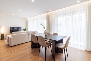 Una sala moderna con sofá, una mesa de comedor con sillas, televisor y un ventanal con cortinas ligeras.