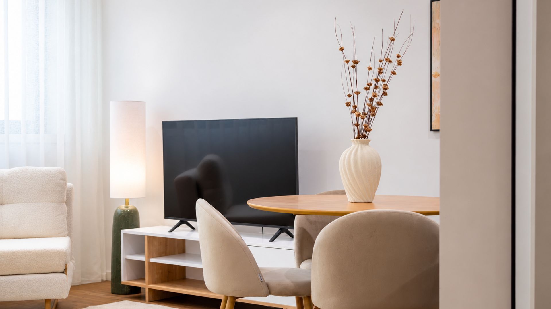 Een moderne woonkamer met een flatscreen televisie, een ronde houten tafel, een beige bank en een witte vaas met gedroogde planten.