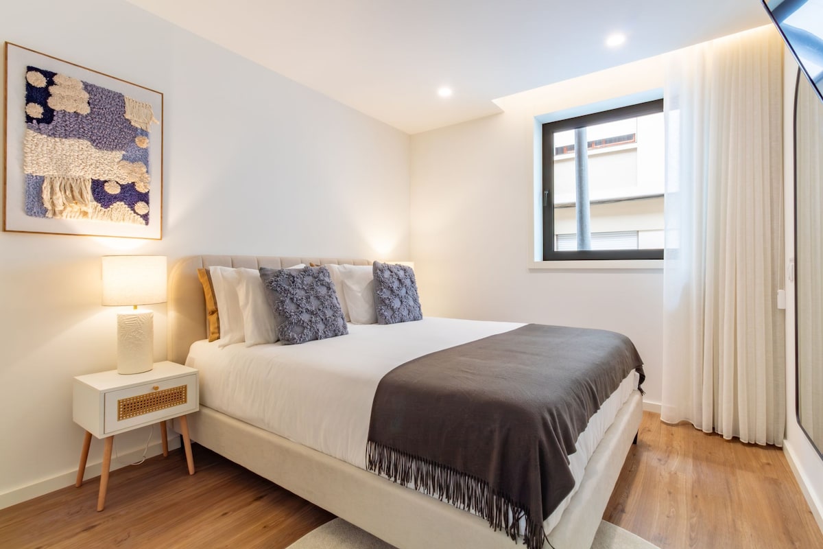 A bedroom with a bed, a bedside table, a lamp, a window with curtains, and a wall-mounted artwork.