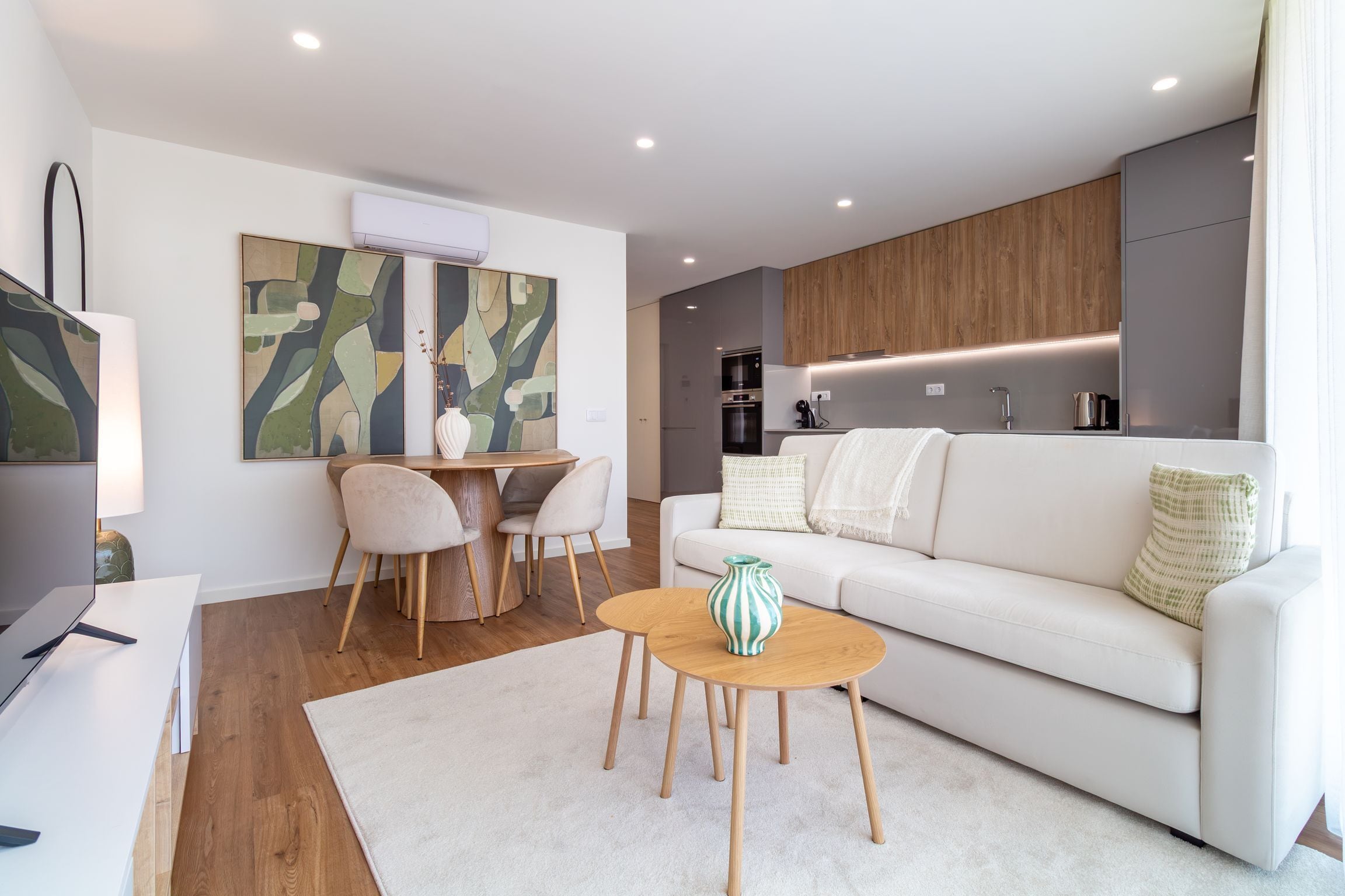 A modern living room with a white sofa, a dining table with chairs, and a kitchen area with wooden cabinets and appliances.
