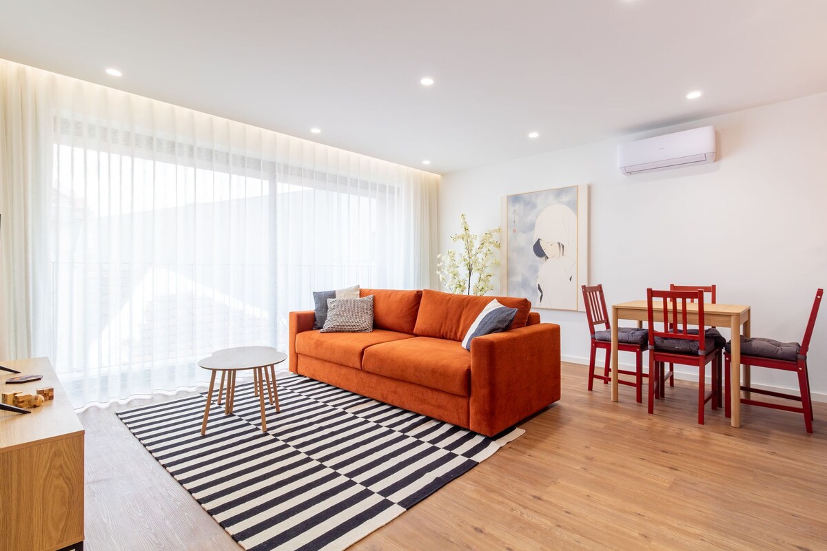 A modern living room with a sofa, a dining table with chairs, and a large window with sheer curtains.