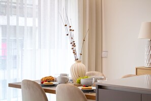Un comedor moderno con una mesa dispuesta para el desayuno, que incluye pasteles, fruta y un jarrón con ramas secas.