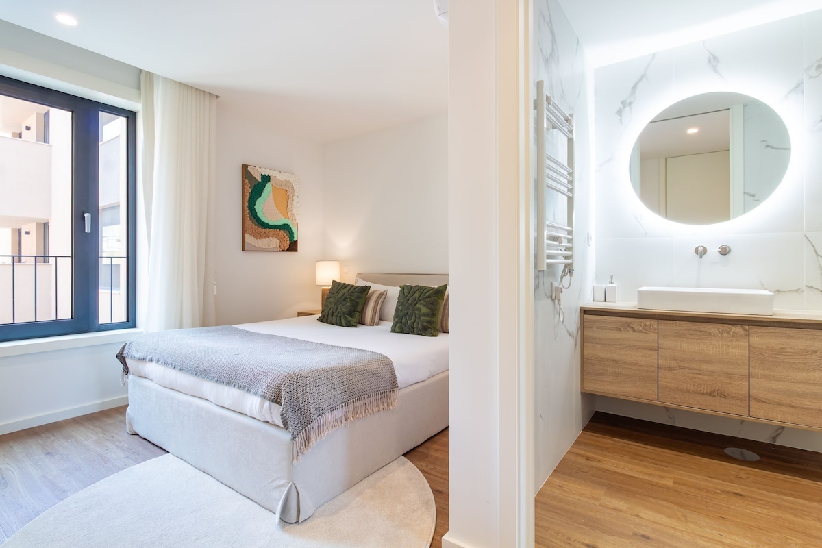 A modern bedroom with a bed, a bedside lamp, a wall-mounted abstract art piece, and a bathroom with a sink and mirror.