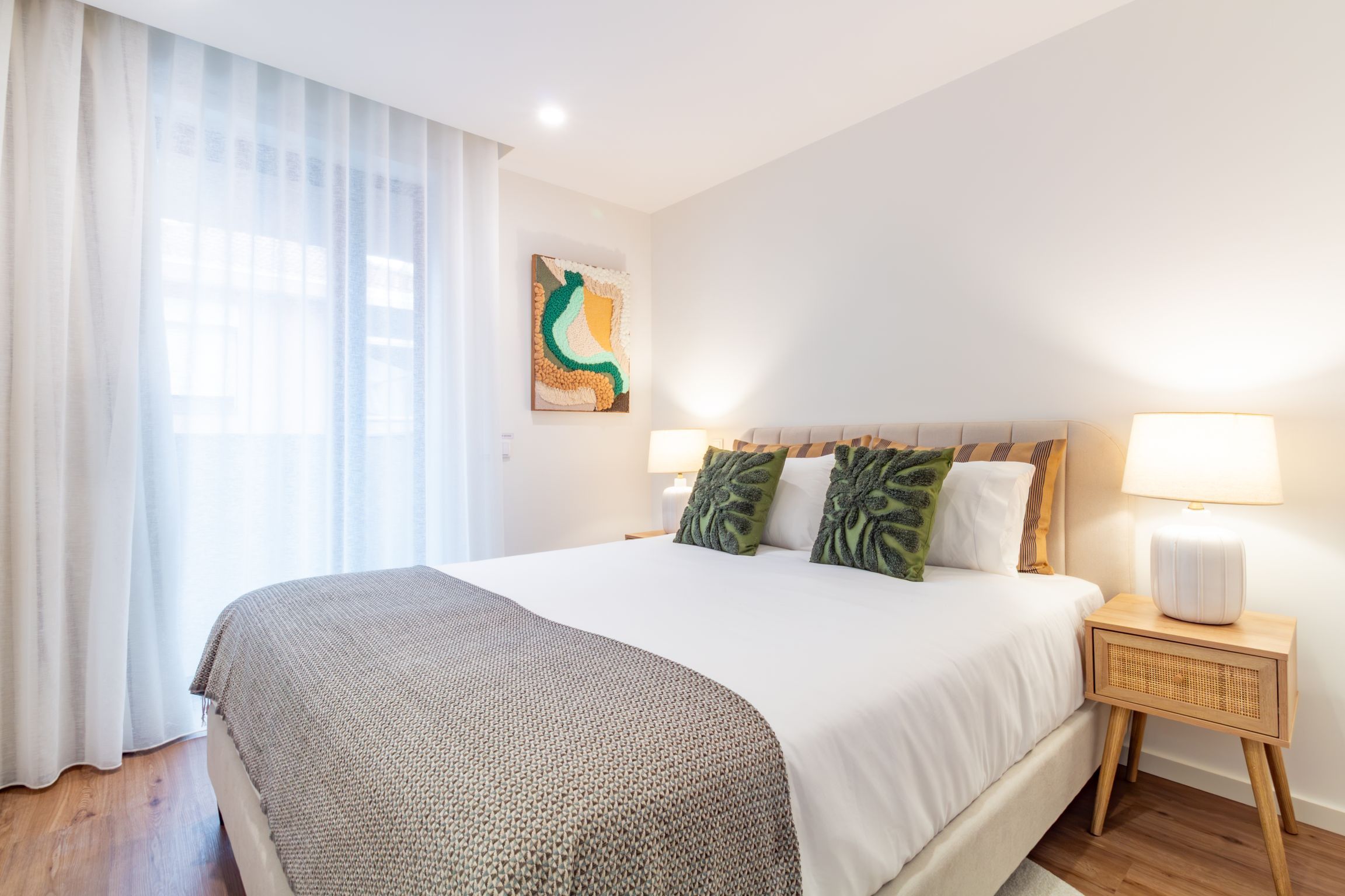 A hotel room with a large bed, a bedside table with a lamp, and a window with sheer curtains.