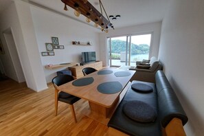 Un espacio habitable moderno con una mesa de comedor de madera, sillas negras, un sofá con cojines, un televisor y vista a un lago.