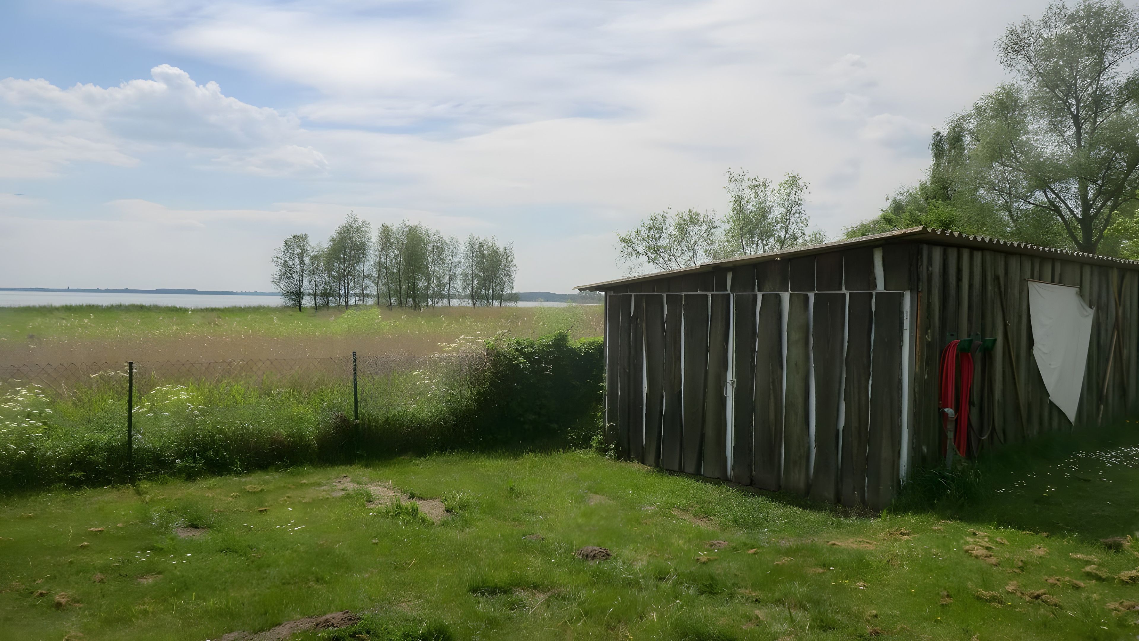 Ländliche Landschaft mit einem Holzschuppen, offenen Feldern und einem entfernten Gewässer.