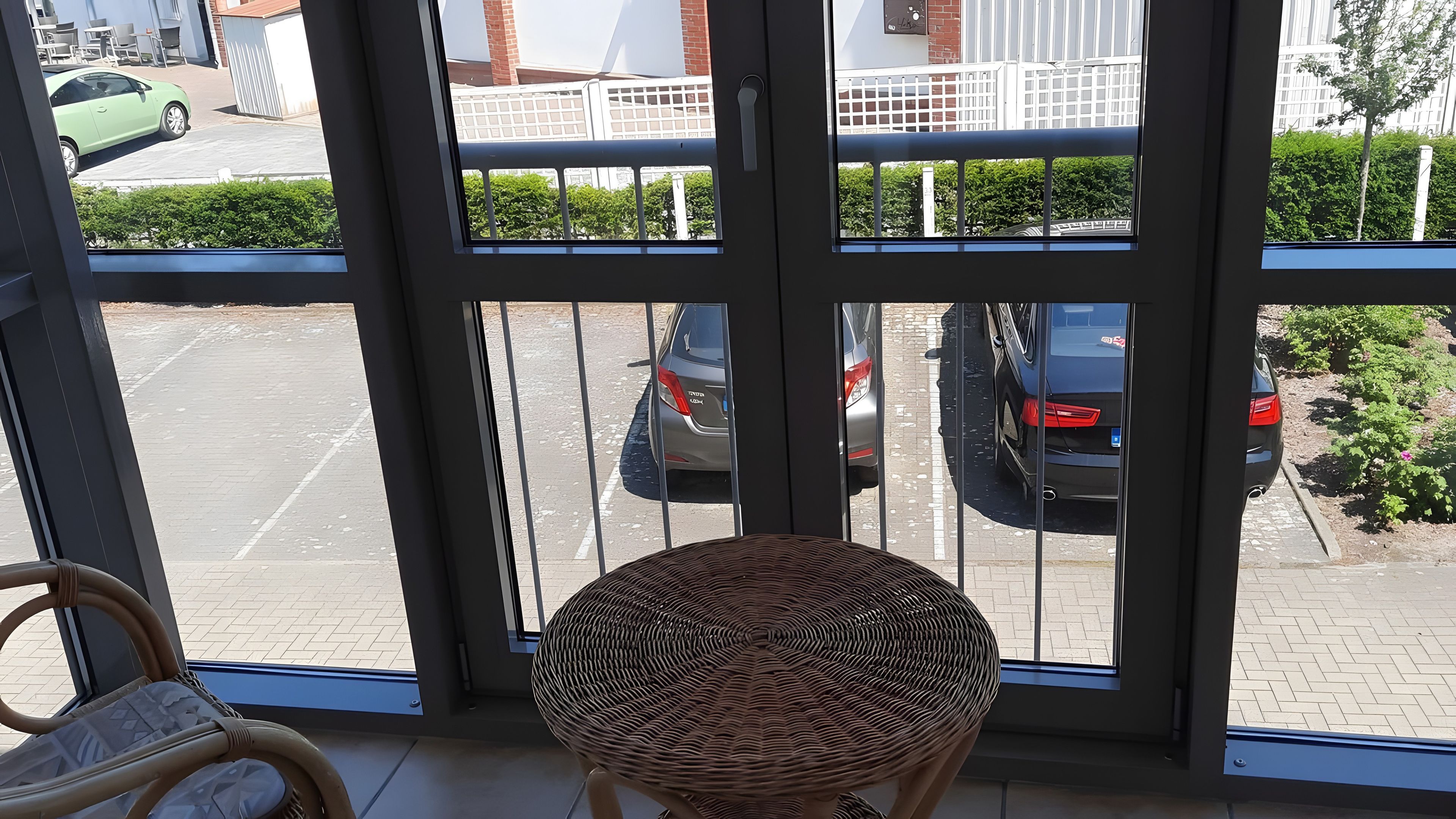 Blick durch eine Glastür, ein Korbstuhl im Innenbereich, ein Parkplatz mit Autos und ein Gebäude mit weißem Zaun im Hintergrund.