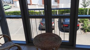Blick durch eine Glastür, ein Korbstuhl im Innenbereich, ein Parkplatz mit Autos und ein Gebäude mit weißem Zaun im Hintergrund.