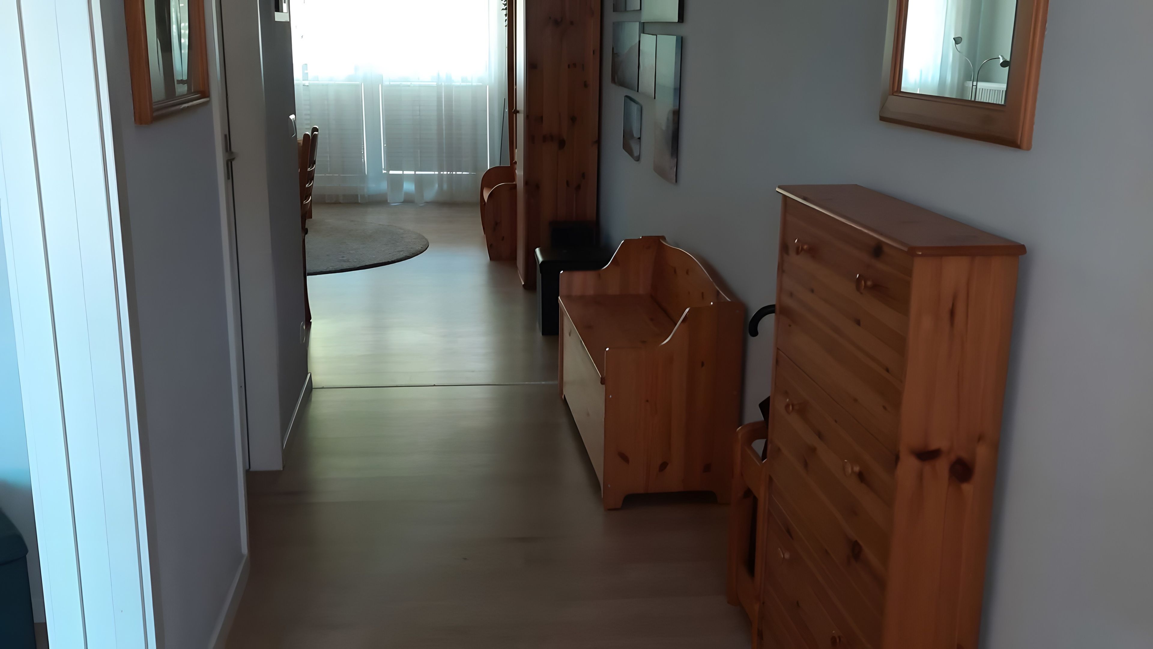 Ein Flur mit Holzmöbeln, darunter ein Schminktisch und eine Bank, der zu einem Raum mit Fenster und Vorhängen führt.
