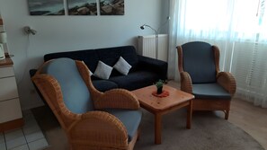 Ein Wohnzimmer mit einem dunkelblauen Sofa, zwei Korbstühlen, einem kleinen Holztisch mit einer Pflanze und einer Heizung.