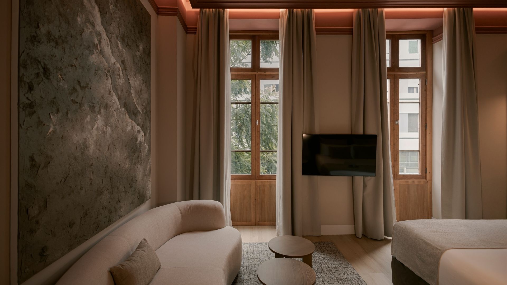 A hotel room with a large stone wall, a sofa, a bed, a TV, and large windows with curtains.