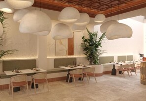 A restaurant with round tables, cushioned chairs, pendant lighting, and a large potted plant.