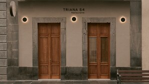 Fachada de edificio con dos puertas de madera, un cartel que indica 'TRIANA 54 Restaurante' y un banco a la derecha.