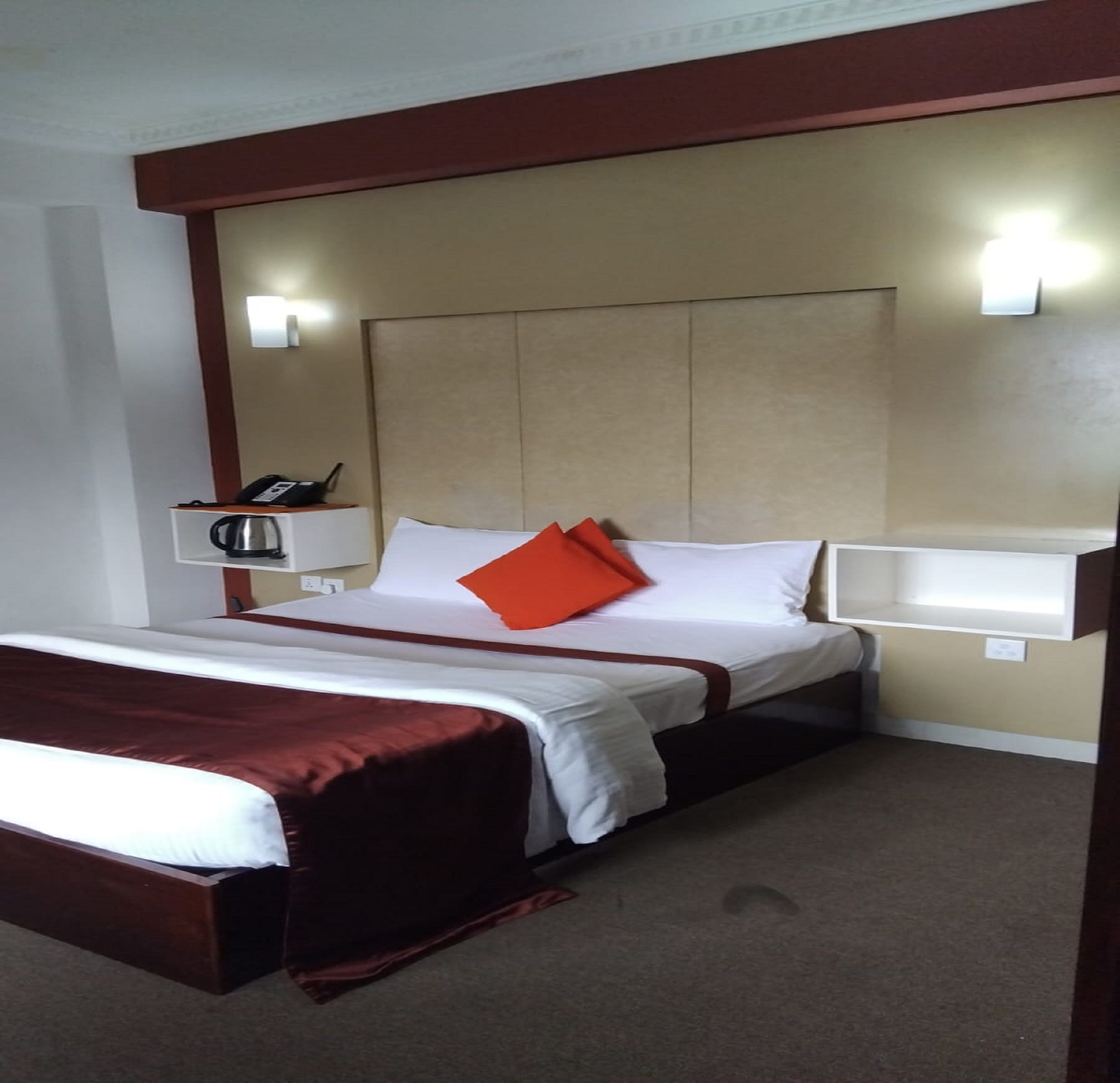 A hotel room with a bed, a bedside table with a phone and a kettle, and a wall-mounted light fixture.