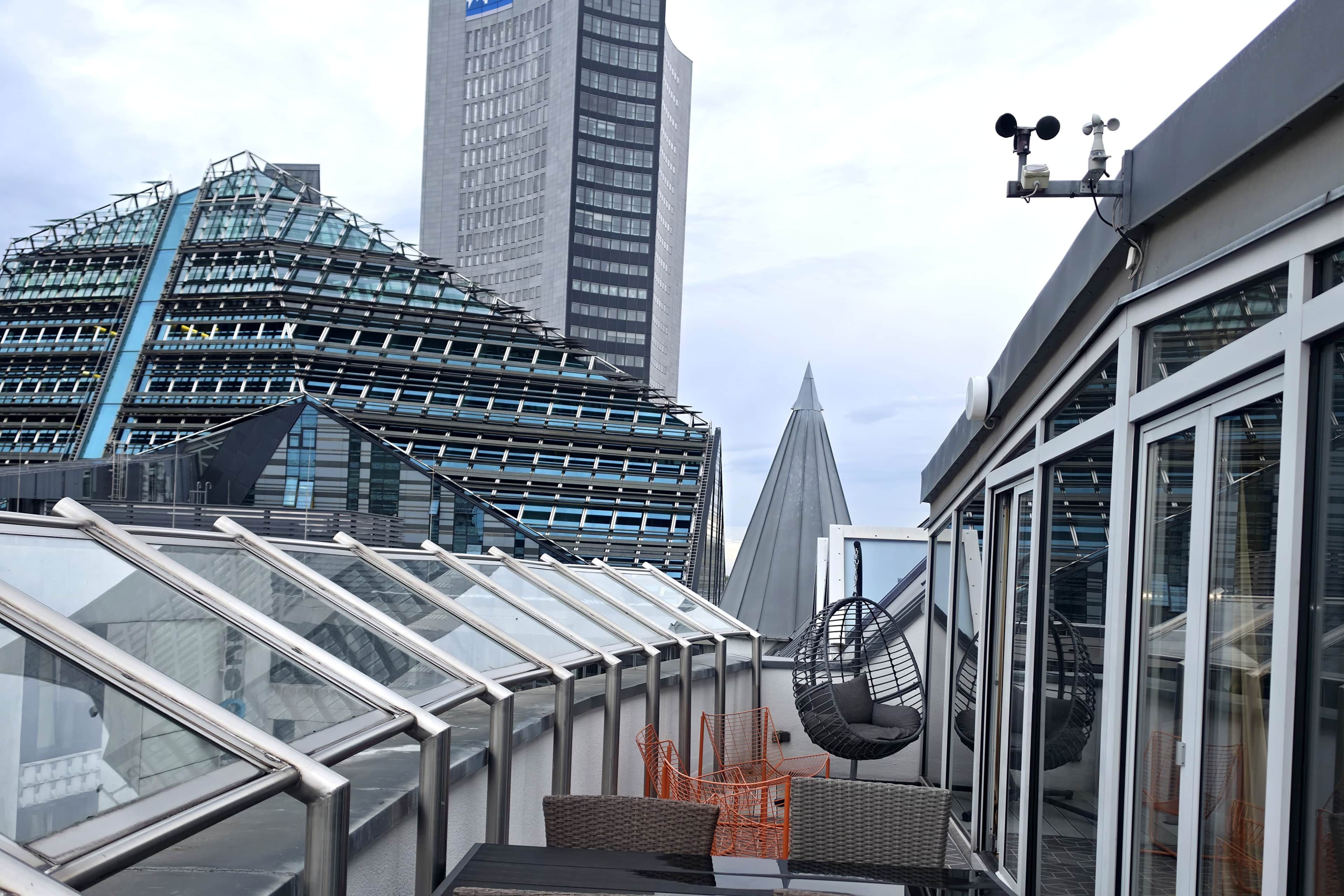 Un bâtiment moderne doté d’une façade en verre, d’une terrasse sur le toit avec des sièges, et offrant une vue sur un gratte-ciel imposant en arrière-plan.