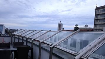 Eine Dachterrasse mit Glasgeländern, Korbmöbeln und Blick auf die Stadtlandschaft.