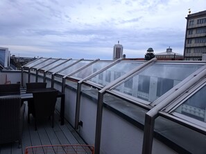 Eine Dachterrasse mit Glasgeländern, Korbmöbeln und Blick auf die Stadtlandschaft.