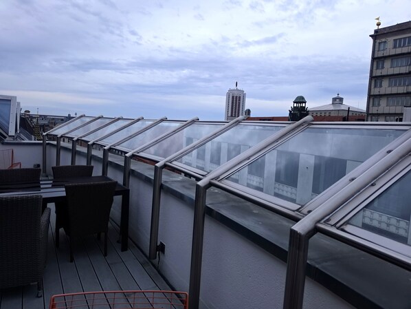 A rooftop terrace with glass railings, wicker furniture, and a view of the cityscape.