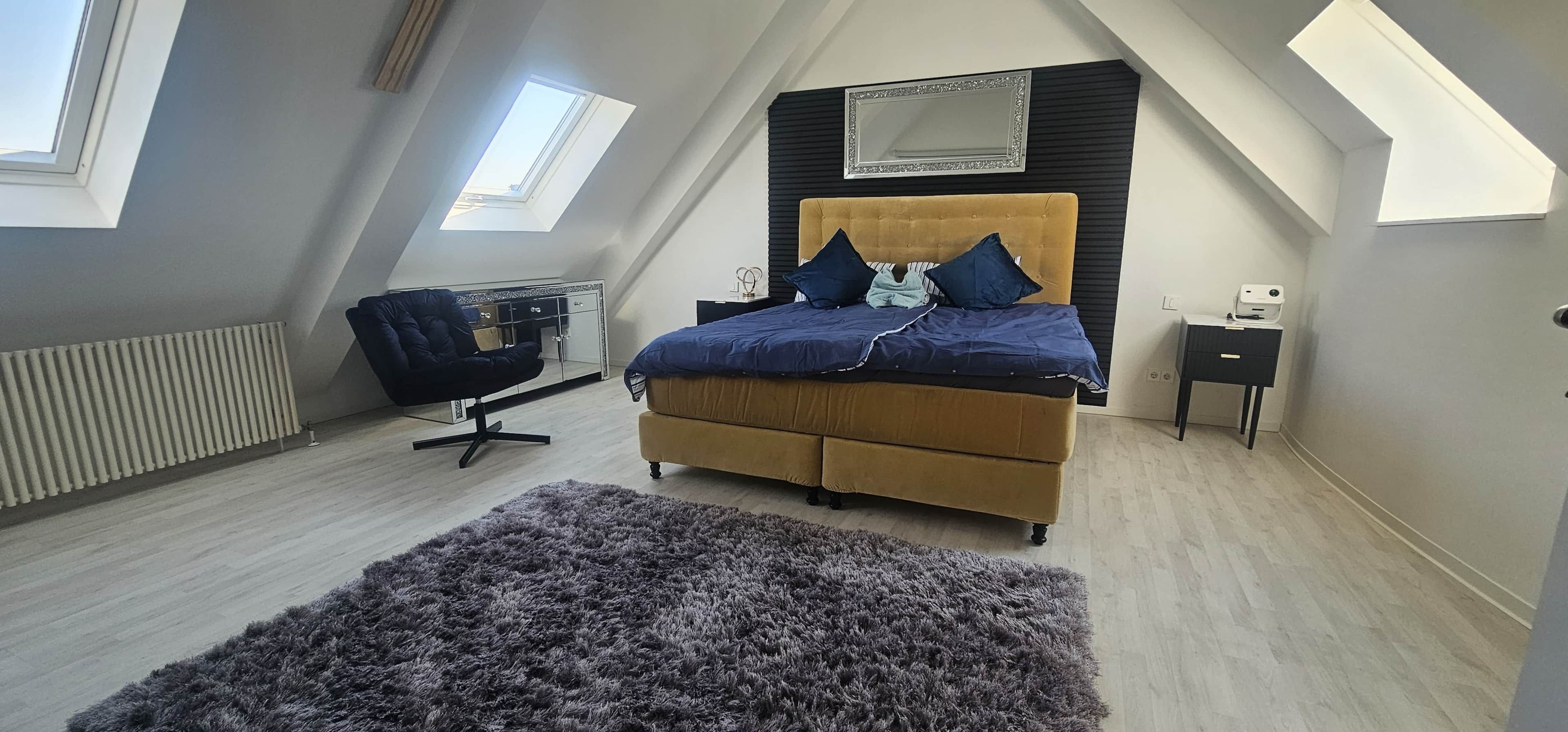A bedroom with a bed, a chair, a small table, a radiator, and a window.