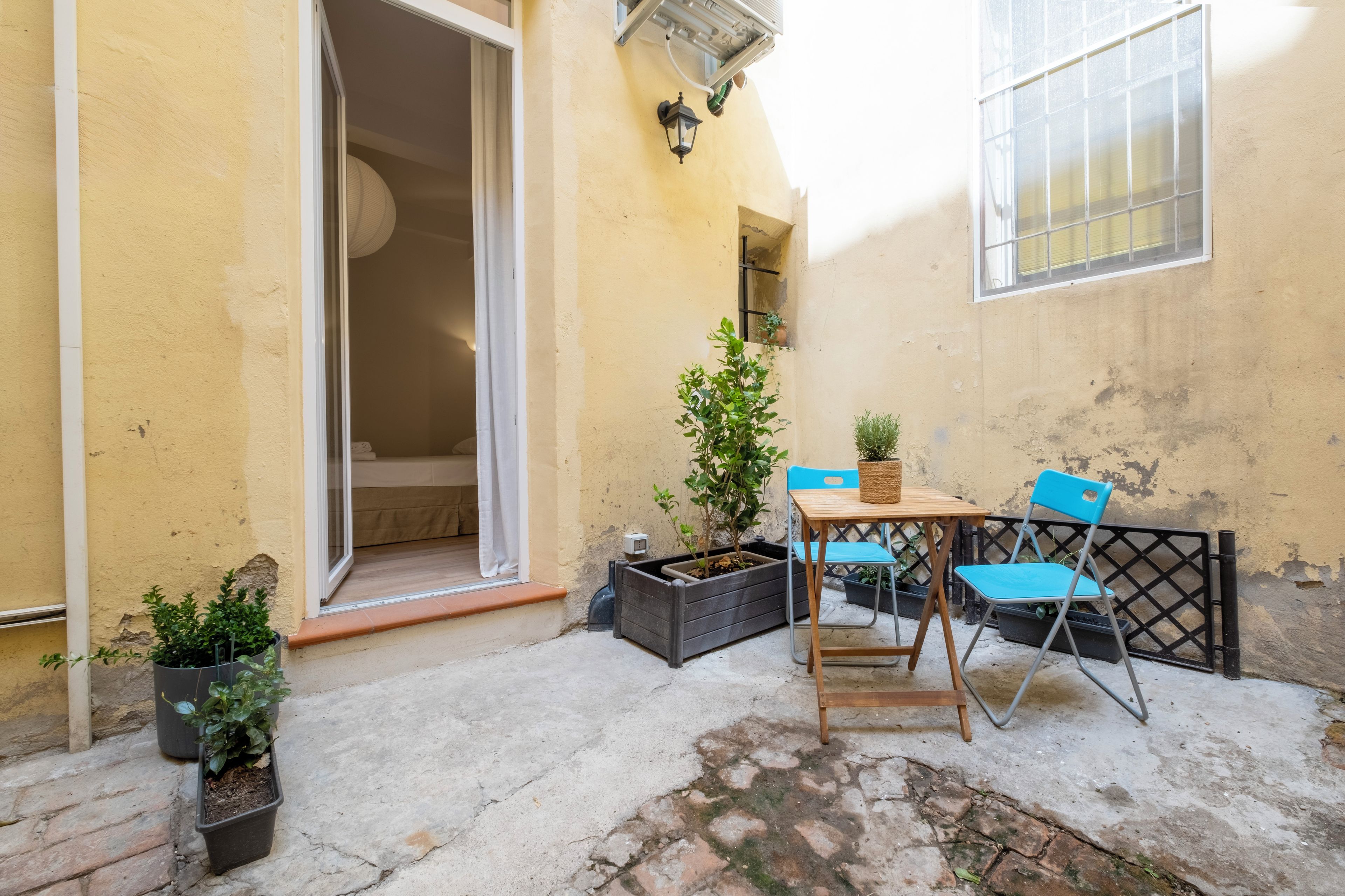 Uma área de pátio externa com mesa, cadeiras, plantas em vasos e uma porta de vidro deslizante que dá para um espaço interior.