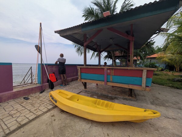 A yellow kayak on a concrete surface, a covered seating area with a bench, and a person standing by a railing. - Serenity Oceanside Guest House (Socorro)