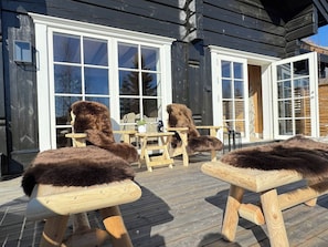An outdoor seating area with wooden furniture and fur-covered cushions, a wooden deck, and a building with large windows and a door. - Familycabin at a Lake (Ringsaker)