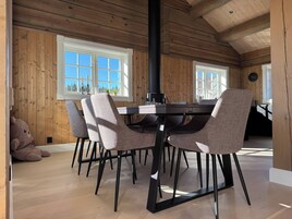 Una habitación con paneles de madera, una mesa de comedor con sillas, un calentador negro y ventanas con vistas a árboles.