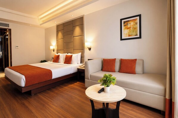 A hotel room with a bed, a sofa, a small table with a plant, and a painting on the wall.