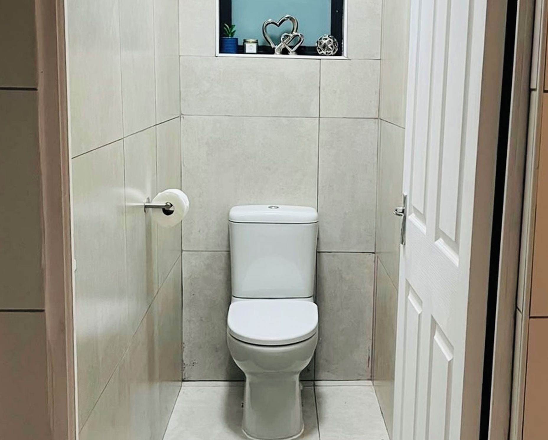 Un baño pequeño y cerrado con inodoro, porta papel higiénico y un estante con objetos decorativos.