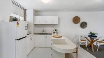 Una cucina compatta con mobili bianchi, un frigorifero integrato, un lavello e un'area pranzo con tavolo e sedie in vetro.