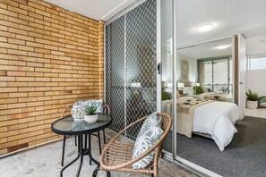 Quarto com cama, uma mesinha com uma planta, uma cadeira de vime e uma porta de vidro deslizante que dá para outro cômodo.