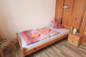 A wooden bed with a floral bedspread, two pillows, a bedside table with a lamp, and a chair with a floral cushion.