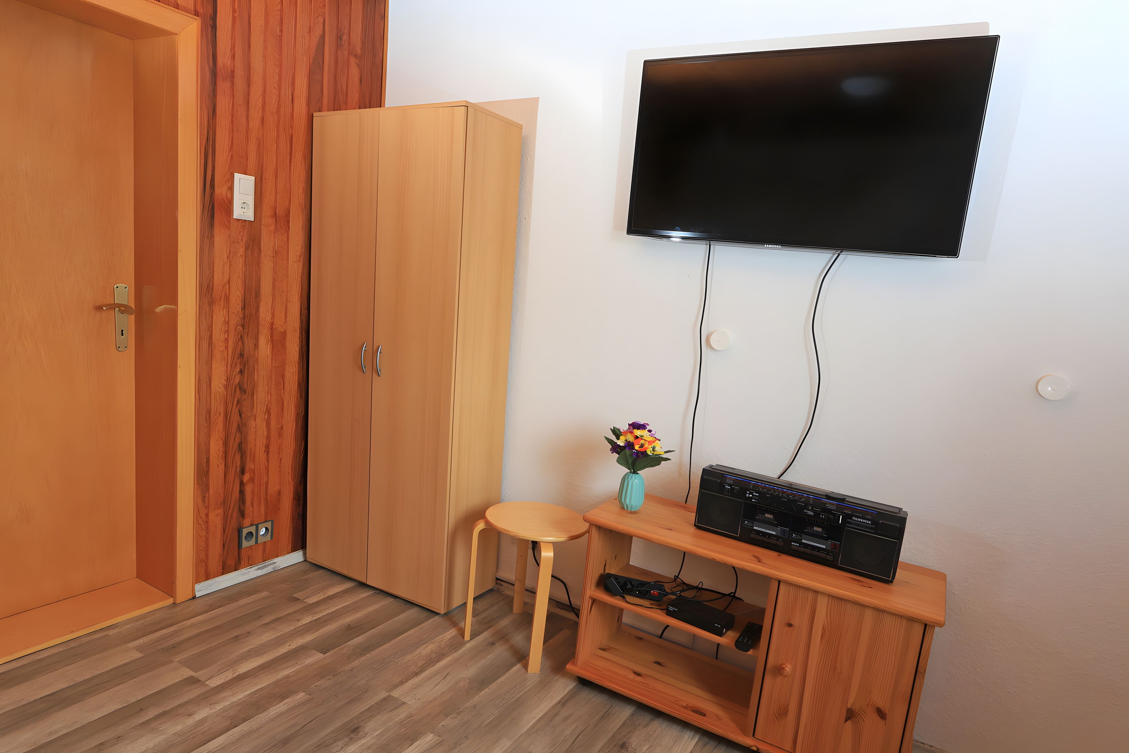Ein Zimmer mit einem Holzschrank, einem an der Wand befestigten Fernseher, einem kleinen Holzschemel und einem Schrank mit elektronischen Geräten.