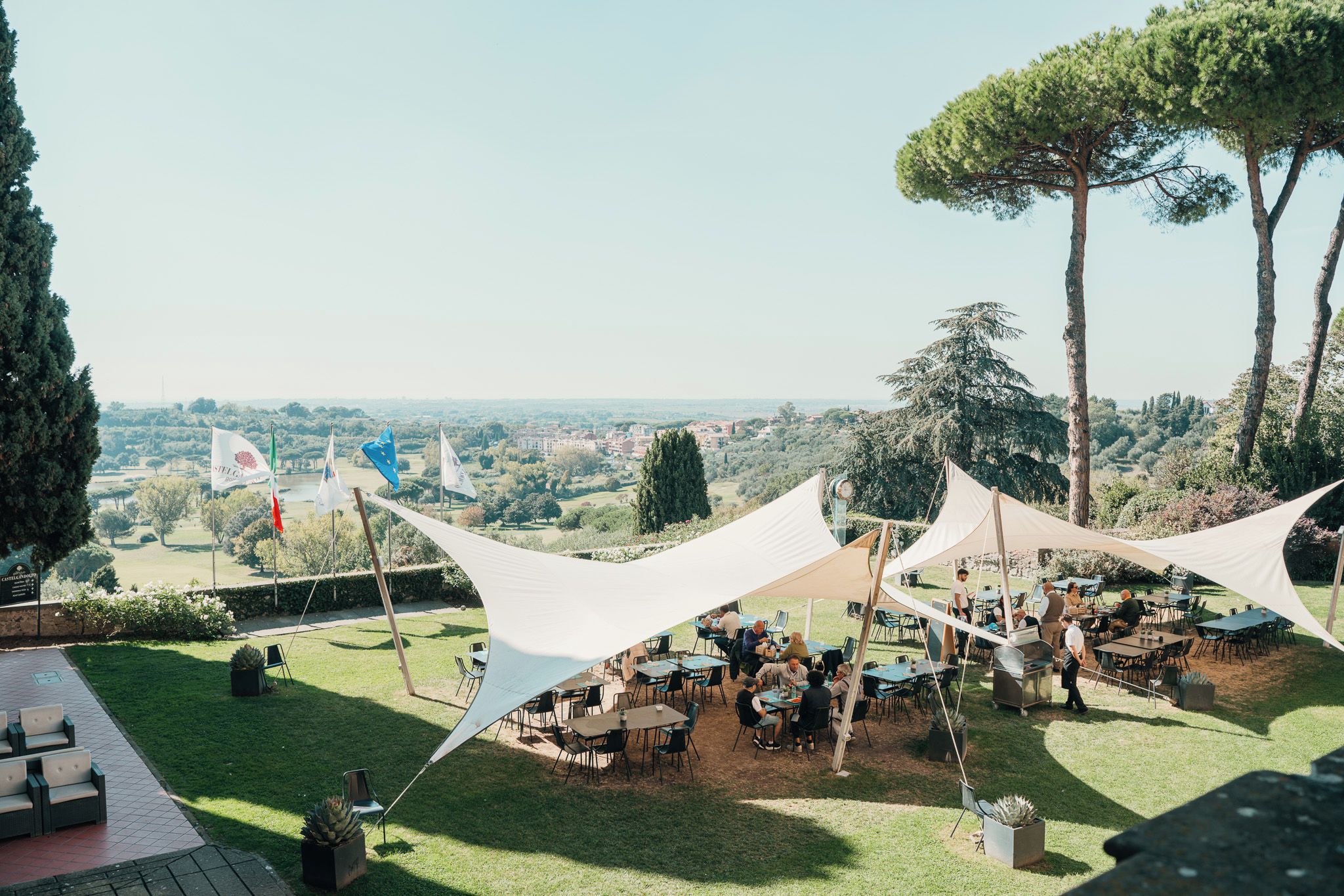 Een evenement buiten met tafels en stoelen onder grote witte tenten, met op de achtergrond bomen en een veraf gelegen stad.