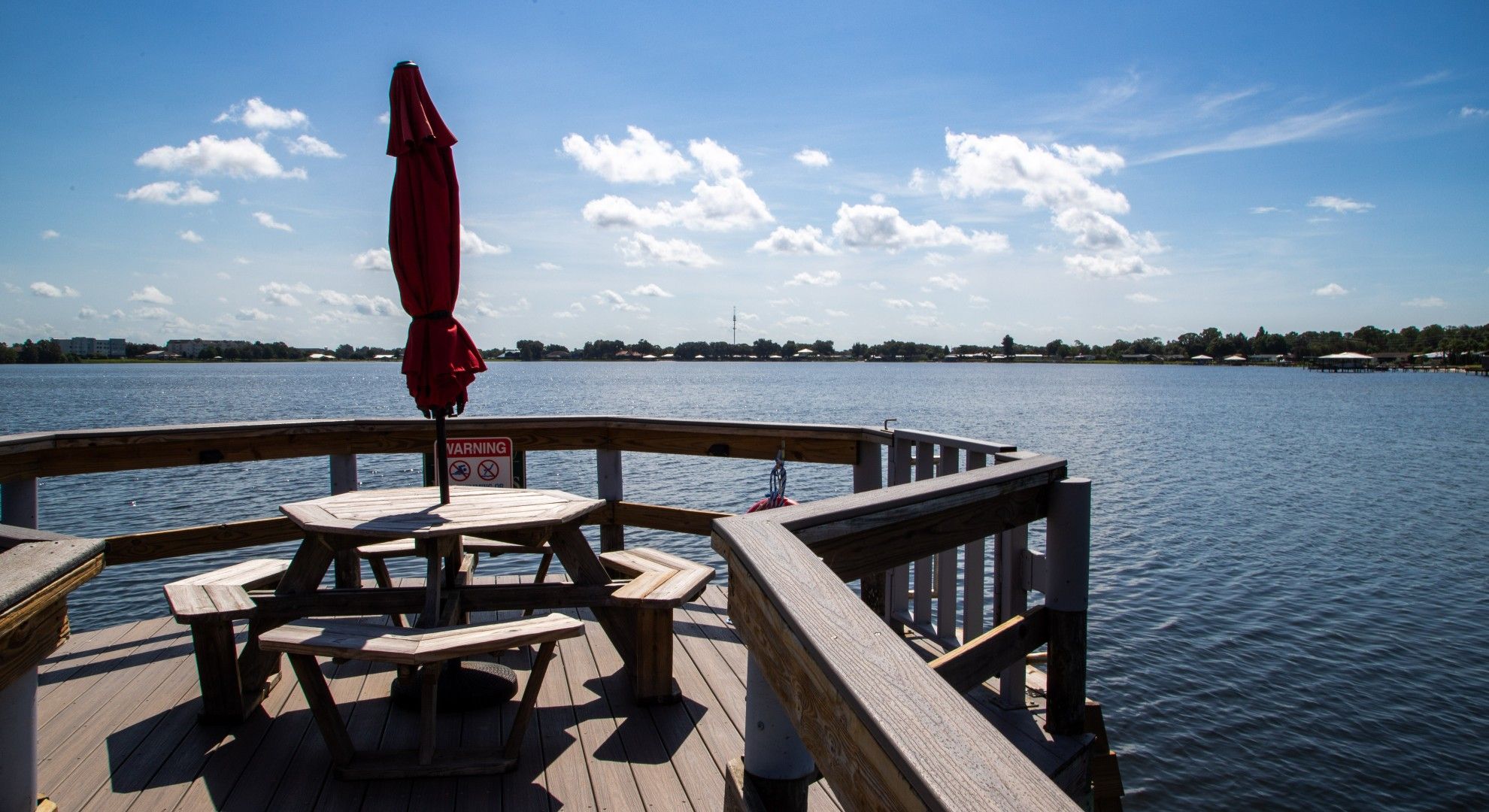 A wooden deck with picnic tables and a red umbrella, overlooking a calm body of water with a distant shoreline.