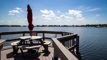 A wooden deck with picnic tables and a red umbrella, overlooking a calm body of water with a distant shoreline.