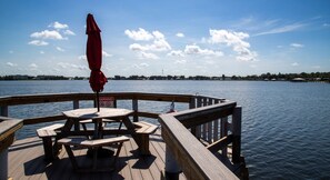 Une terrasse en bois, dotée de tables de pique-nique et d’un parasol rouge, surplombe une étendue d’eau calme dont on aperçoit la rive au loin.