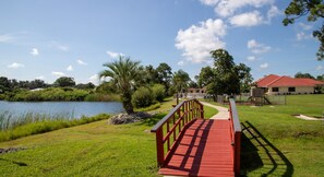 Un pont en bois mène à une zone herbeuse où se trouve un plan d’eau et un bâtiment surmonté d’un toit rouge.