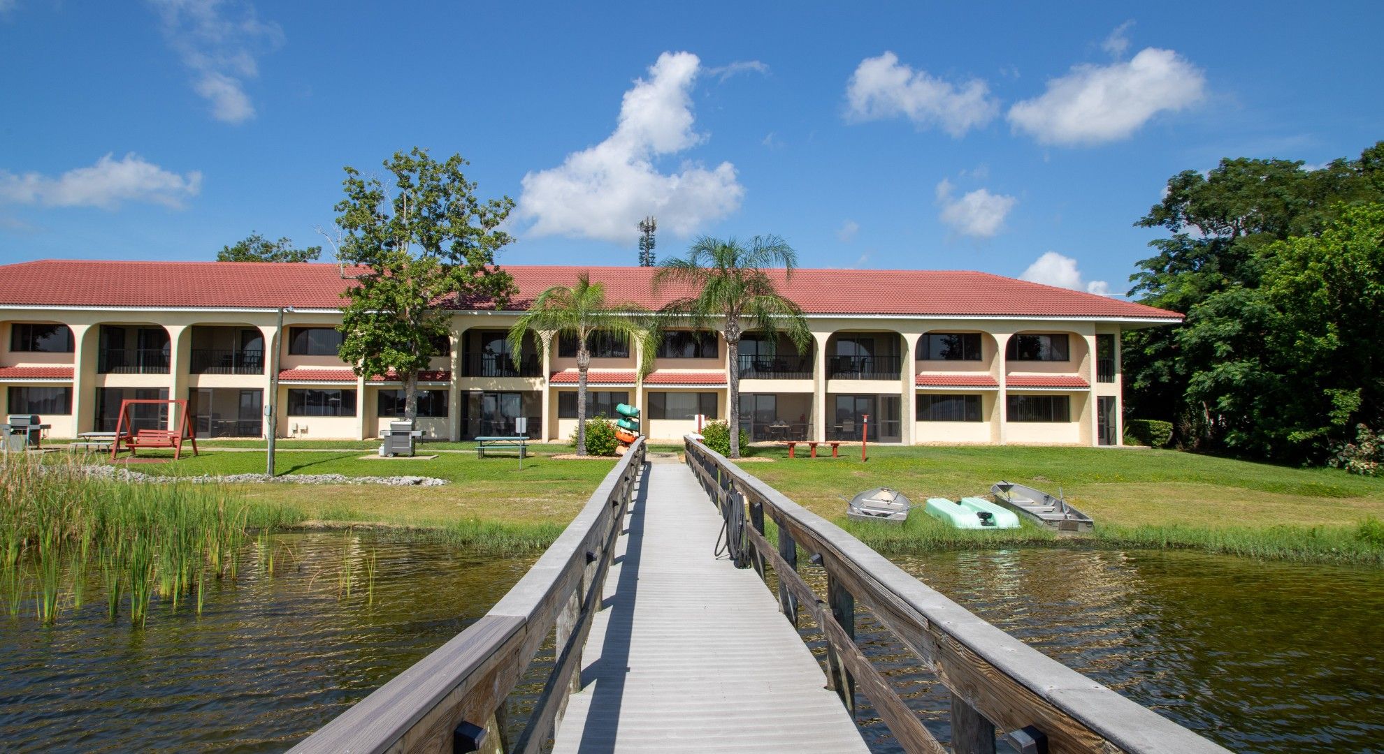 A hotel with a red-tiled roof, a wooden dock leading to a body of water, and a grassy area with trees.