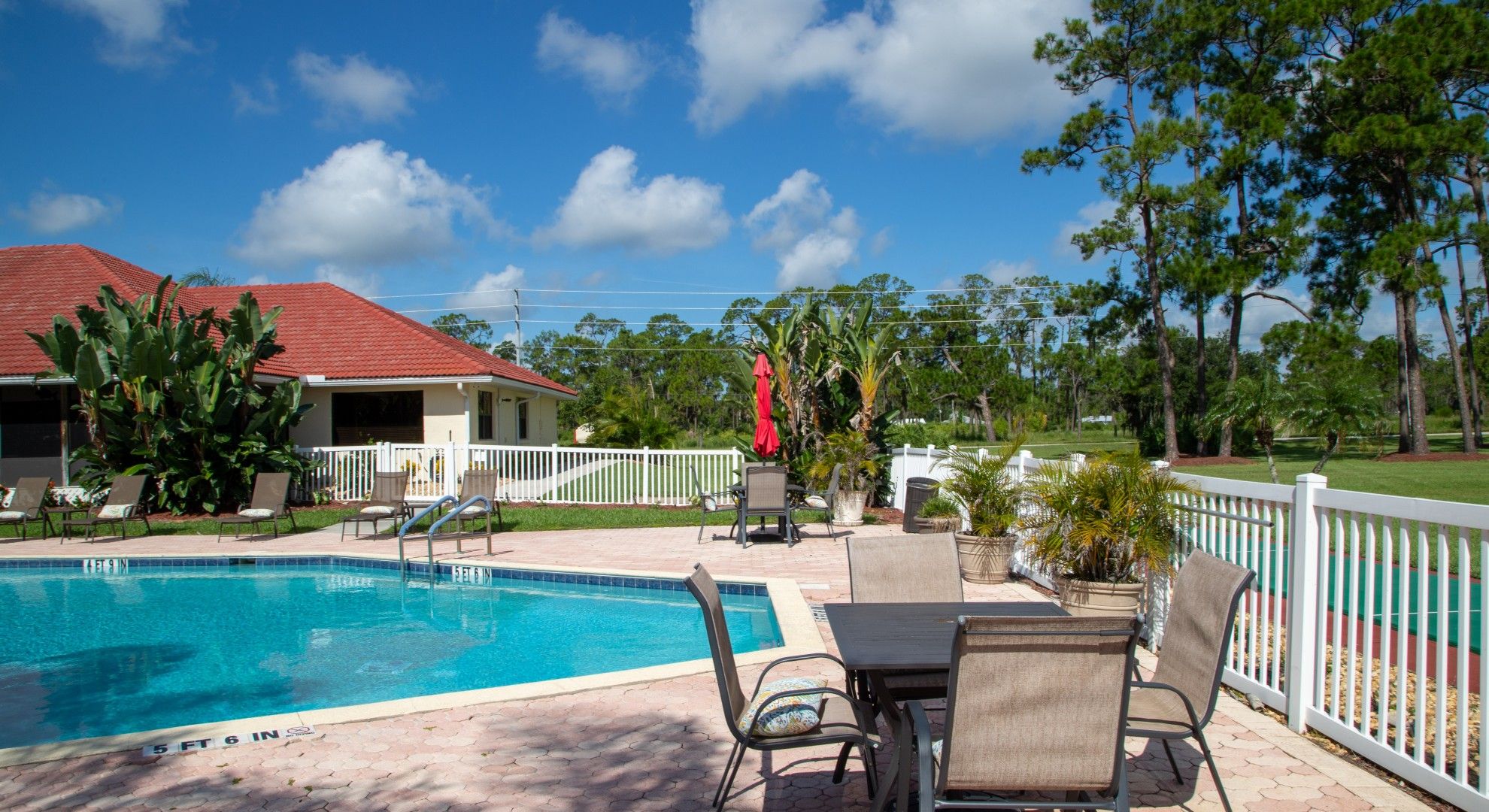 A swimming pool with lounge chairs and a table, surrounded by a white fence and trees.