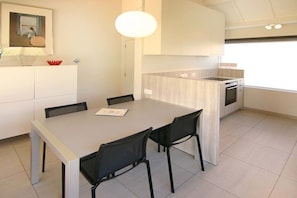 A modern kitchen with a dining area, featuring a light-colored table and chairs, a pendant light, and a view of the outside. - Holiday apartment with 1 bedroom (Cadzand-Bad)