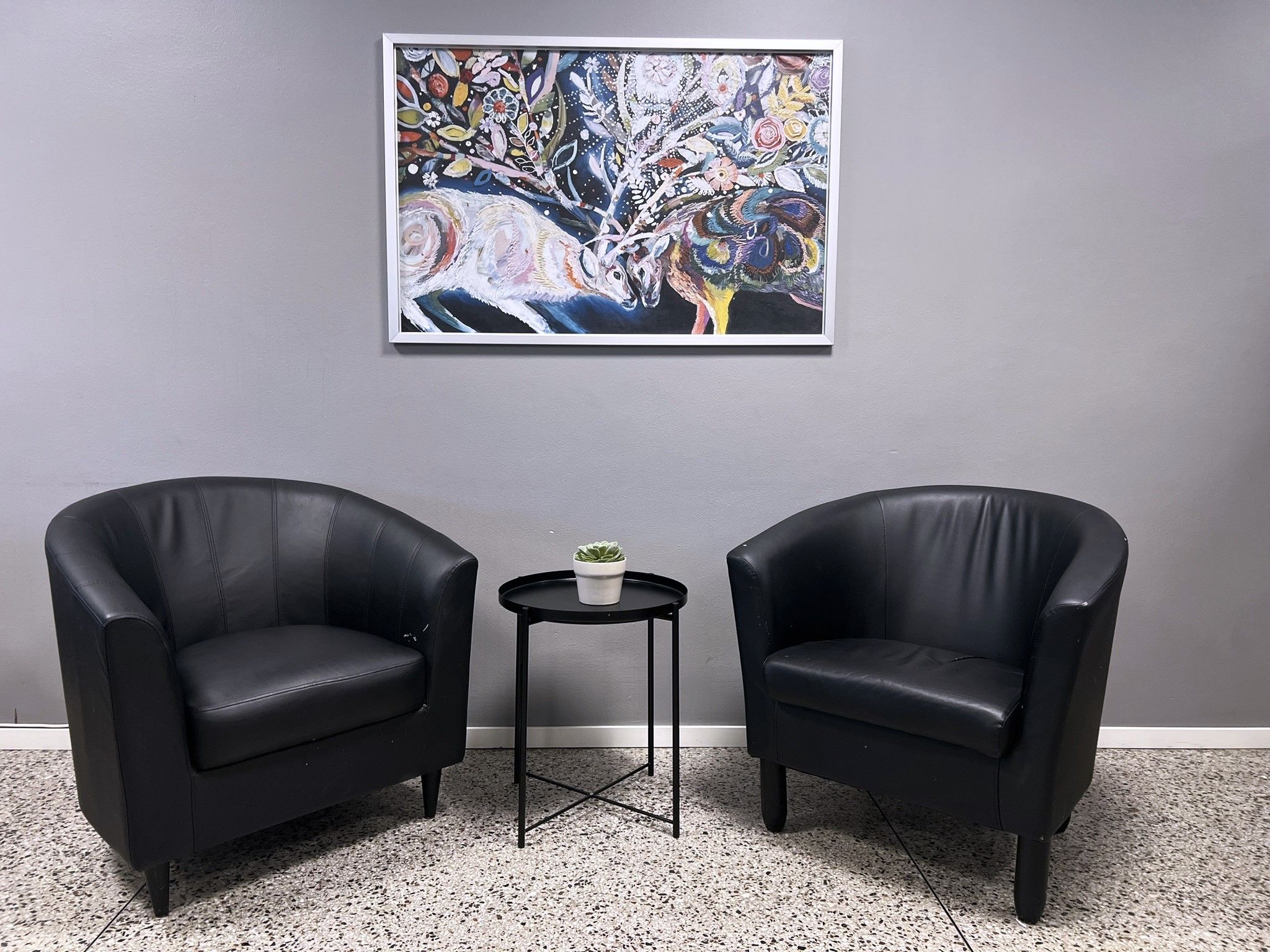 Duas poltronas de couro preto, uma mesa redonda pequena com uma planta em vaso e um quadro abstrato colorido na parede.