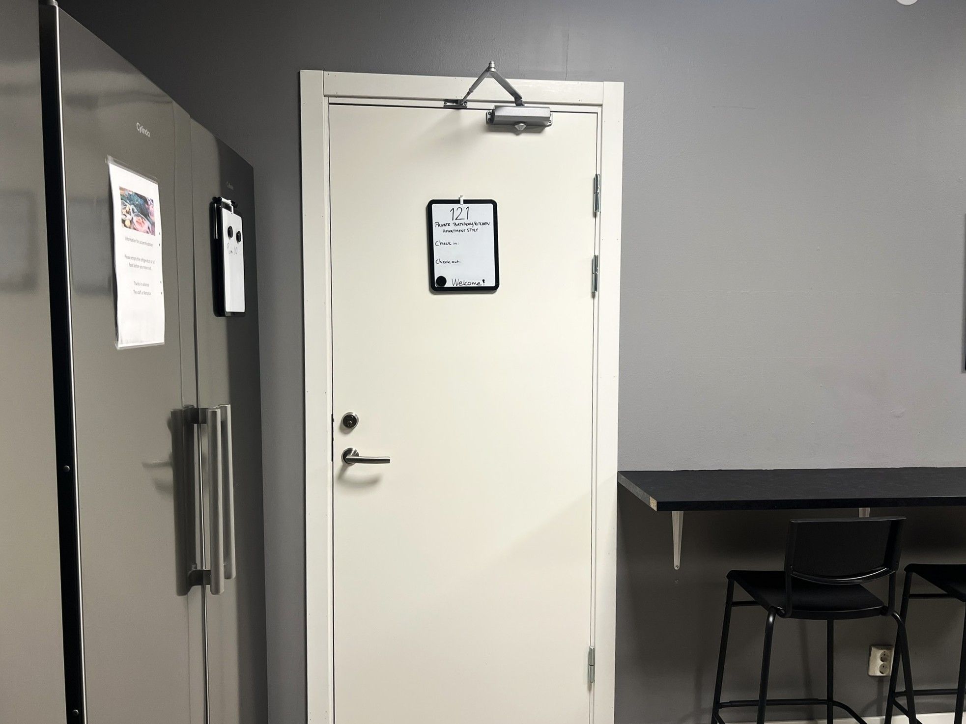 A door with a sign indicating a private bathroom and kitchen, a desk with a chair, and a wall-mounted light fixture.