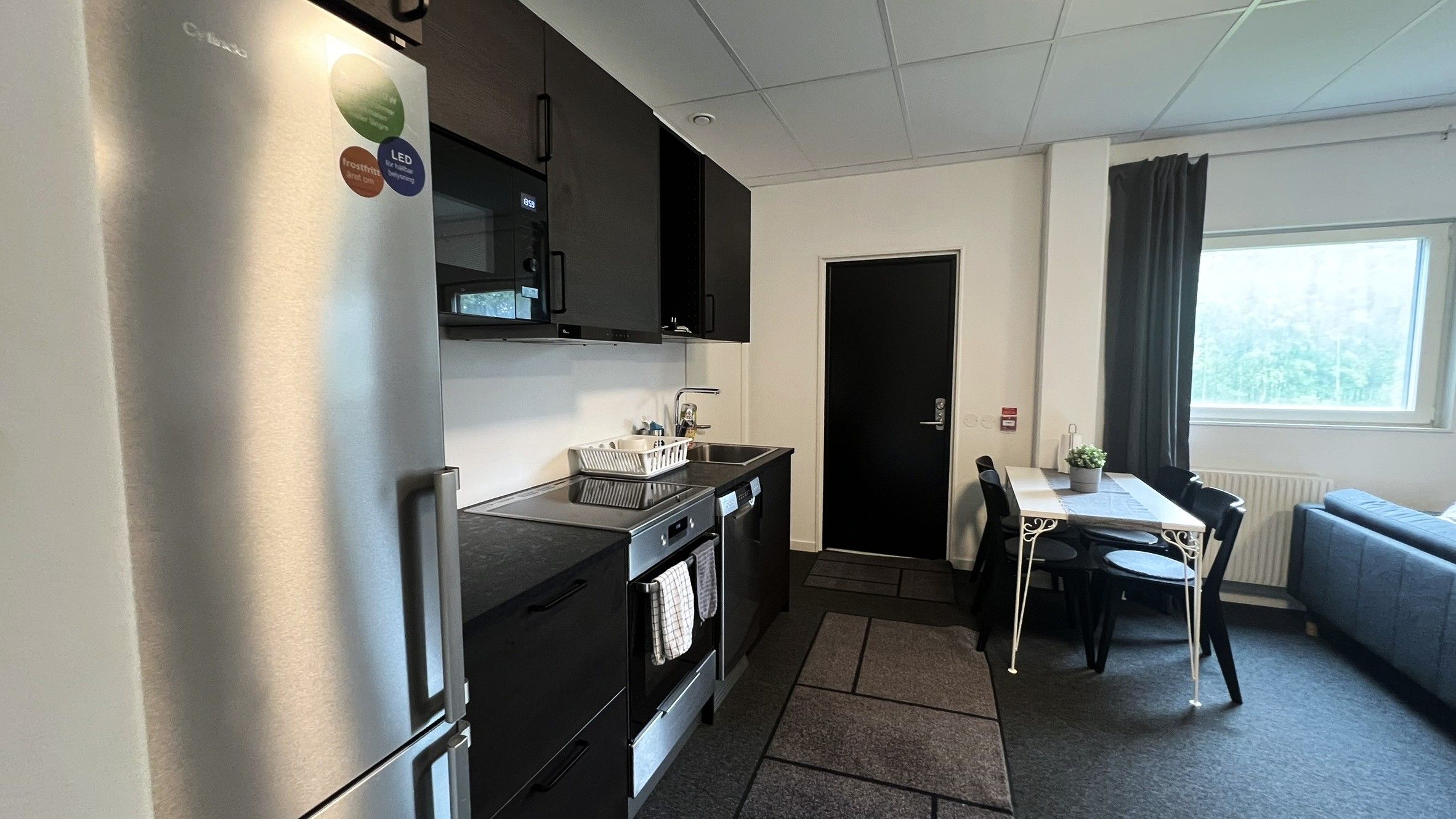 A compact kitchen with a refrigerator, stove, and sink. There's a dining area with a table and chairs, and a window with curtains.