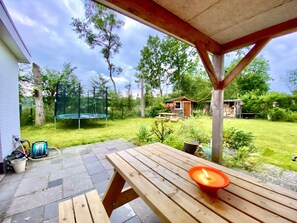 Een houten terras met een tafel en een kaars, een trampoline en een klein houten bouwwerk in een achtertuin.