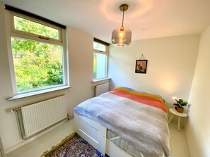 Ein Schlafzimmer mit einem Bett, einem Heizkörper, einem Fenster, einer Hängeleuchte und einem kleinen Tisch mit einer Pflanze.