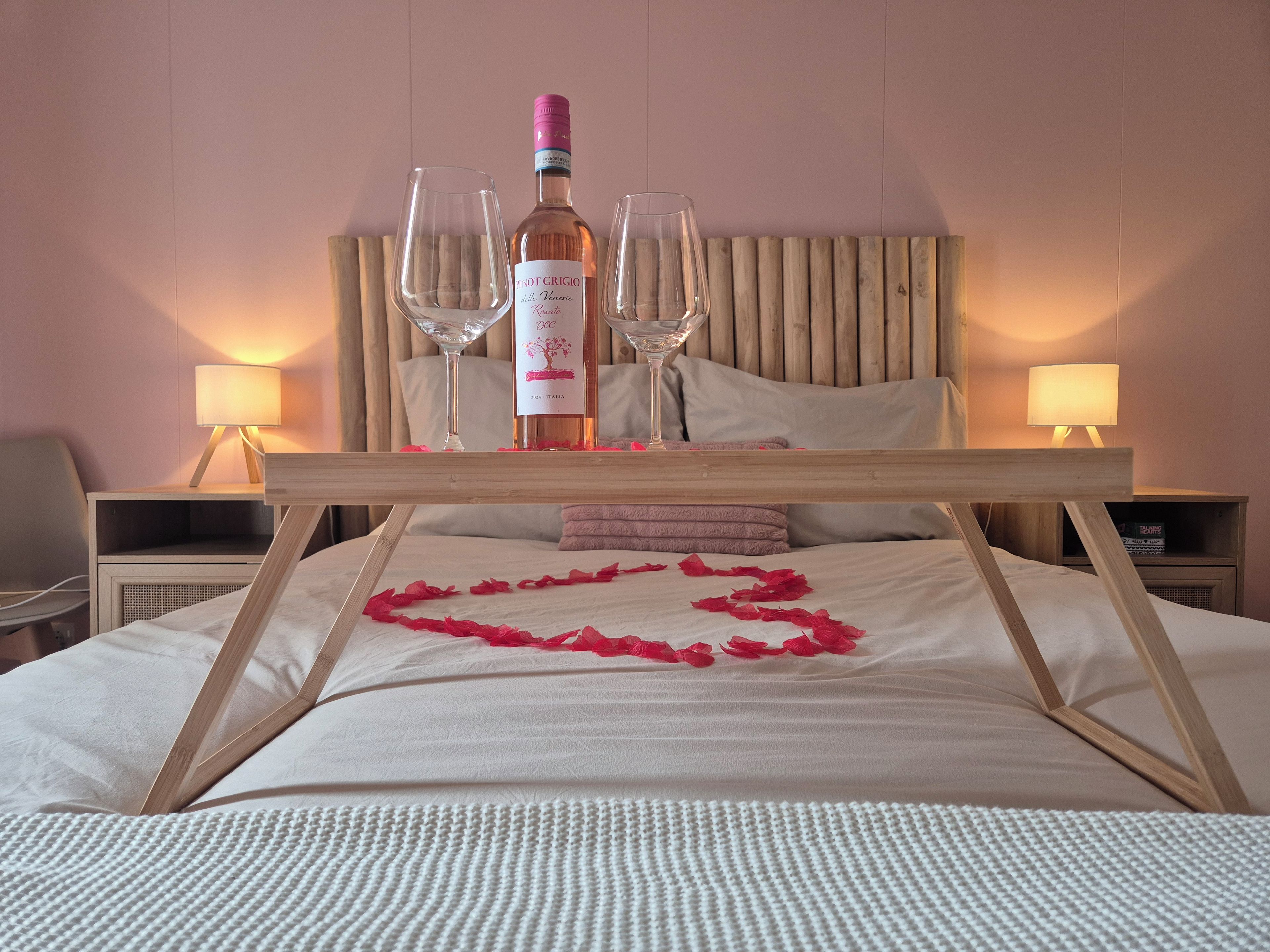 A bed with a wooden frame, a bottle of wine, two glasses, and a heart-shaped decoration made of rose petals.